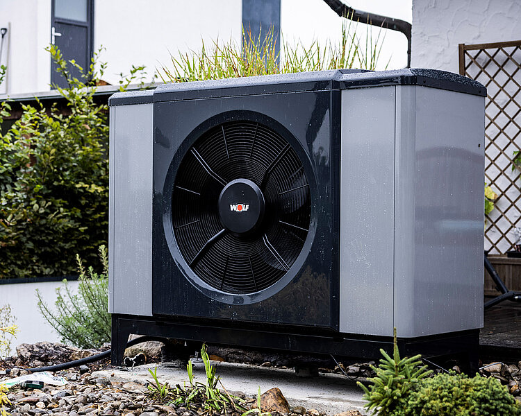 WOLF Wärmepumpe im Außenbereich installiert Außen aufgestellte WOLF Wärmepumpe in modernem Gartenbereich mit Kies und Bepflanzung vor Wohnhaus