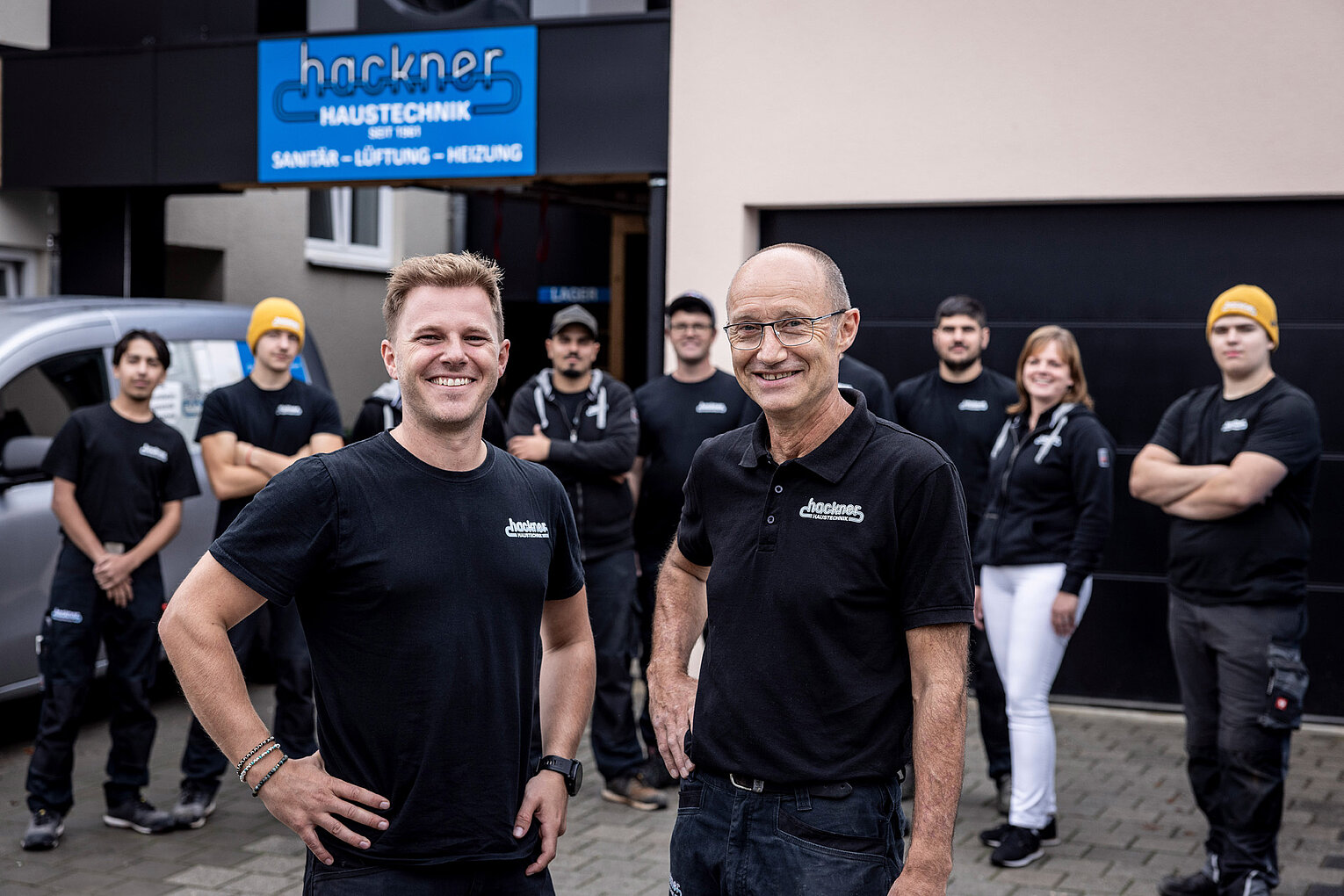 Geschäftsführung mit Team von Hackner Haustechnik Zwei Führungskräfte von Hackner Haustechnik lächeln in die Kamera, das Team steht im Hintergrund